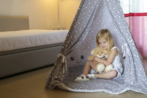 Cute toddler girl sitting in wigwam with her teddy bear in bedroom