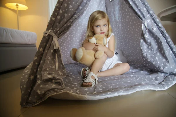 Cute toddler girl sitting in wigwam with her teddy bear in bedroom