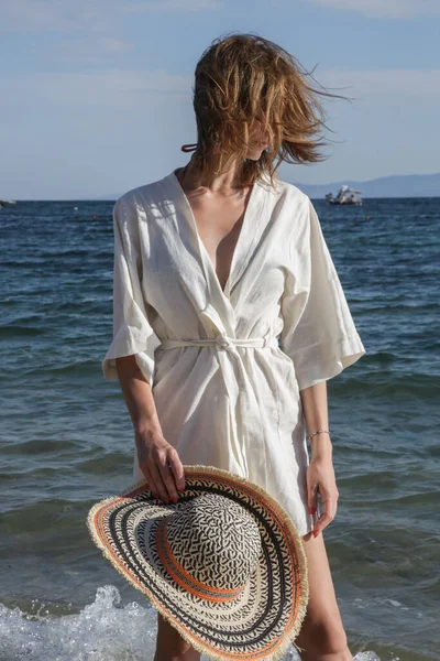 Plajda keten kimono elbise ve büyük hasır şapka giyen genç bir kadının portresi.