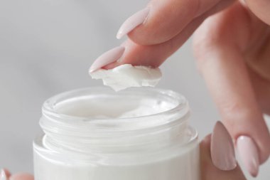 Kadının elinin yakınında, parmağıyla kavanozdan krema alıyor. Temiz ve yumuşak cilt için doğal malzemeler içeren kozmetik ürünler kullanılıyor. Sağlık, cilt bakımı ve güzellik konsepti.