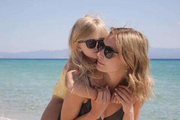 Mother carrying daughter on her back on the beach. Happy idyllic family summer vacation concept.