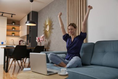 Oturma odasında dizüstü bilgisayarı üzerinde çalışırken kollarını kaldıran mutlu bir kadın.