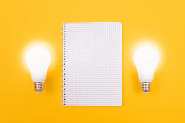 Glödande energisparlampa med anteckningsblock liggande på gult bord — Stockfoto