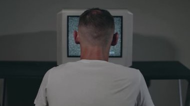 Kırık Televizyon ya da TV Propagandası - Beyaz tişörtlü Zombi Adam Karanlık Oda 'da Eski Gümüş TV' ye Karşı Oturuyor. Televizyon sinyalini ara. Ekranda bozulma. Geri Vizyon, Statik Çekim, Film Tanesi