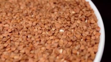 Dry Uncooked Brown Buckwheat Groats on White Plate Rotating against Black Background. Raw Large Buckwheat Grains. Russian, Ukrainian and Belarusian Culture