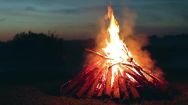 Big Burning Campfire in Early Morning or Evening against the Blue Sky. Wood on Fire. Flying Sparks. Travel and Tourism Concept. Giant Flaming Bonfire at Summer - Static Shot, Slow Motion
