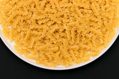 Pişmemiş Fusilli Makarna Siyah Arkaplanda Beyaz Tabakta Yatıyor. Çiğ ve kuru makarna. Sağlıksız ve Yağlı Gıda - Üst Görünüm