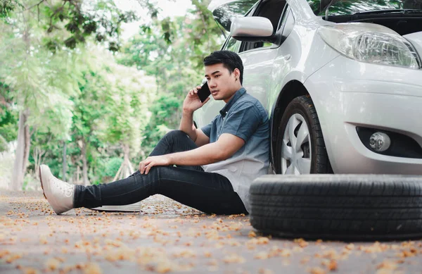 アジア人男性が車の横に座り 路上で車が故障した後 携帯電話を使って支援を求めている 車両エンジンの問題や事故や専門の機械工からの緊急支援の概念 — ストック写真