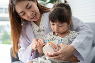 Asyalı kadın diş hekimi, diş fırçası ve stomatologla dişlerini fırçalayan tatlı küçük bir kıza diş sağlığı kliniğinde diş sağlığı, eğitim ve önleme boşlukları kavramını anlatıyor..