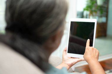 Üst düzey Asyalı kadın gözlük takıyor ve evinde kanepede dinleniyor ve haber ve e-kitap okumak için tablet kullanıyor. Yaşlılar için teknoloji kavramı.