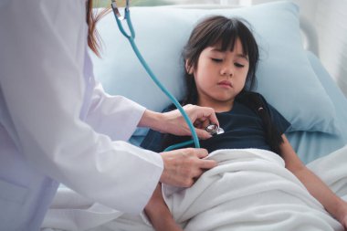 Asyalı kadın pediyatrist doktor muayene için stetoskop tutuyor küçük bir kız hasta ve çocuğun kalp ciğerleri, iyi aile doktoru çocuğu evde ziyaret ediyor, sağlık ve çocukluk konsepti için ilaç alıyor..