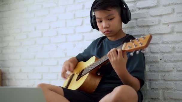 アジアの少年は オンラインのためのラップトップとビデオ会議で友人や教師とオンラインで音楽を再生するための仮想会議でギターを演奏することを学ぶ インターネット上のコミュニケーション学習コンセプト — ストック動画