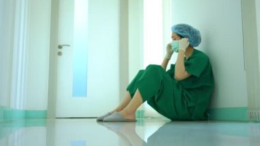 Stresli ve çok çalışan kadın doktor doktor, hastane odasının yanında yerde oturuyor ve maskeyi çıkarıyor.
