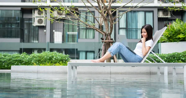 Mutlu genç girişimci kadın havuz kenarında solaryum yatağında oturuyor ve bilgisayarını uzaktan kumandalı dijital iş projesi için kullanıyor.