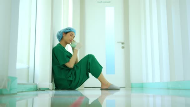 病院の手術室の近くの床の上に座ってマスクを取るストレスと過労の女性医師医療従事者の外科医の側面図 — ストック動画