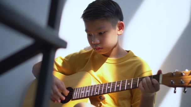 自宅のリビングルームで居心地の良いソファに座って座っているアコースティックギターを演奏し 好きな趣味 音楽教育の概念イメージを楽しむアジアの少年の前に — ストック動画