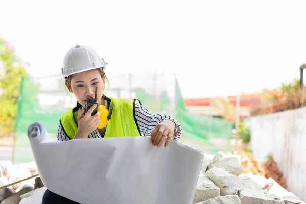 Asian engineer or Young woman Architect put on a helmet for safety and look at Blueprint for Inspect Building factory Construction Site and use walkie talkie for a talk with a contractor.
