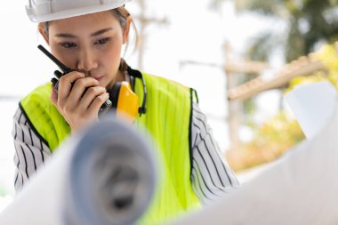 Asyalı mühendis ya da genç kadın mimar güvenlik için bir kask taktı ve İnşaat Fabrikası İnşaat Alanının Şablonuna baktı ve müteahhitle konuşmak için telsiz kullandı..
