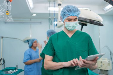 Tıbbi maskeli Asyalı cerrahın portresi ayakta duruyor ve bir hastanede ameliyathanede tablet tutuyor. Profesyonel cerrahlar takımı. Sağlık hizmeti, acil tıbbi servis kavramı