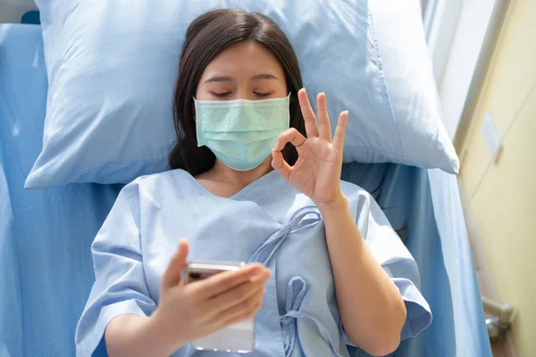 Happy Asian female patient smiling, lies on the bed, and Shows an ok symbol to show confidence in treatment. Concept of believe in treatment And insurance coverage