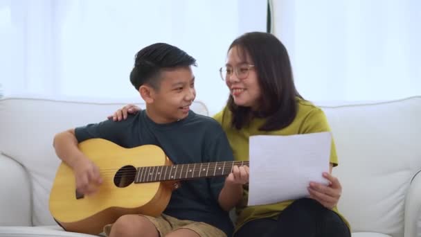 亚洲母亲拥抱着儿子 亚洲男孩弹吉他 母亲抱在沙发上 感受到赞赏和鼓励 幸福家庭的概念 学习和快乐的生活方式 爱的家庭关系 — 图库视频影像