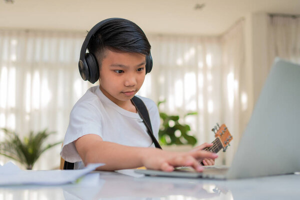 Asian boy playing guitar and watching online course on laptop while practicing for learning music or musical instrument online at home. Boy students study online with video call teachers play guitar.