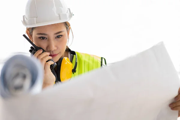 Asian engineer or Young woman Architect put on a helmet for safety and look at Blueprint for Inspect Building factory Construction Site and use walkie talkie for a talk with a contractor.