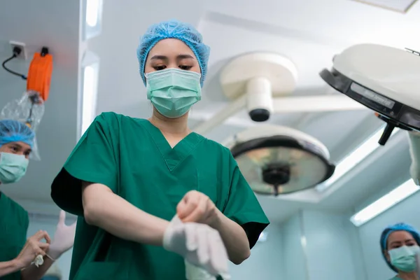 Portrait of Asian surgeon with medical mask standing and wearing medical gloves in operation theater at a hospital. Team of Professional surgeons. Healthcare, emergency medical service concept