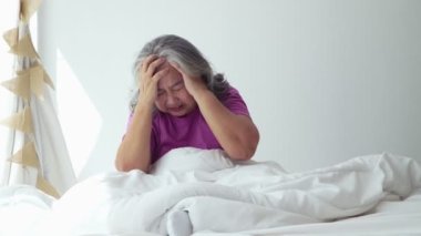 Senior woman with gray hair in bed feel depressed or suffering from strong headache migraine and high blood pressure, sitting alone. Migraine Elderly Lonely sad old woman hold her head With Hands
