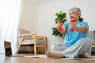 Asian senior woman lifting dumbbell for exercise and workout at home. Active mature woman doing stretching exercise in living room. Exercise Active and healthy for older, elder, and senior concept.