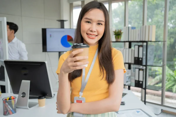 Young Asian Happy Businesswoman or intern holding coffice cup in Modern Office background is Colleagues discuss with new startup project Idea. Empowered Digital Entrepreneur Works on Startup Project