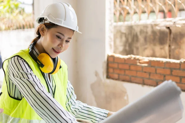 Asian engineer or Young woman Architect put on a helmet for safety and look at Blueprint for Inspect Building factory Construction Site and use walkie talkie for a talk with a contractor.