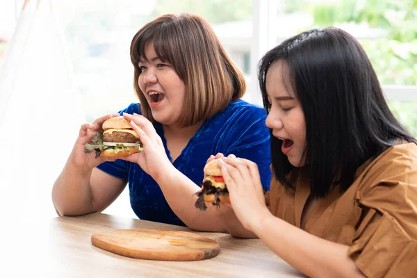 Hungry overweight woman holding hamburger on a wooden plate, During work from home, gain weight problem. Concept of binge eating disorder (BED).