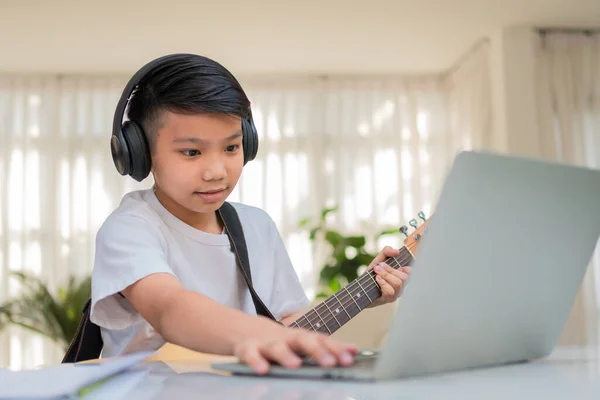 Asian boy playing guitar and watching online course on laptop while practicing for learning music or musical instrument online at home. Boy students study online with video call teachers play guitar.