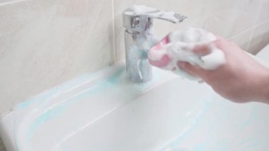 Bir el, sünger kullanarak köpüklü deterjanı lavabonun ve banyo musluğunun üzerine iyice sürer. Gidonu ve musluğu yıkıyorum.