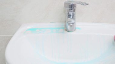 Lastik eldivensiz pembe süngerli bir el banyodaki beyaz lavaboyu ve musluğu iyice yıkar.