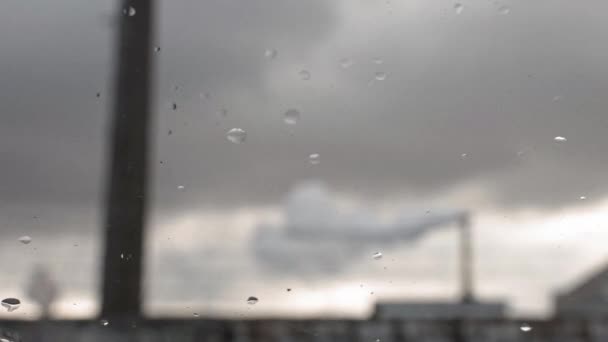 Fokus pada latar depan kaca dengan tetesan hujan. Asap berasal dari cerobong asap di latar belakang terhadap langit yang gelap dan bengkak — Stok Video