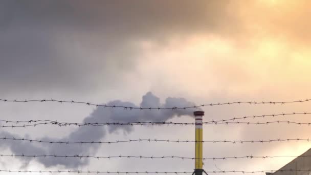 Smoke comes from a yellow chimney against the sky, barbed wire in the foreground — Stockvideo