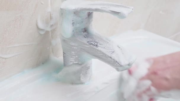Een hand met een roze spons zonder rubberen handschoenen wast de witte gootsteen en kraan grondig in de badkamer — Stockvideo