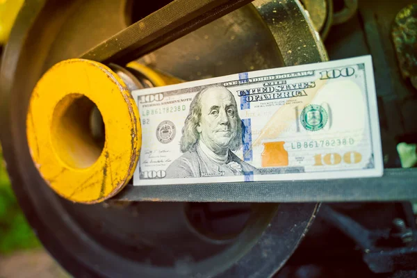 Hundred dollar bill close-up on an industrial background. Money on a belt driven tractor engine pulley. Production costs and the financial component of big business