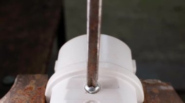 A Phillips screwdriver unscrews a screw from a white power plug close-up. The process of parsing the body of a plastic fork clamped in a vice. Repair and maintenance of electrical equipment. High