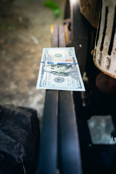 A hundred dollar bill lies on the engine drive belts of a rural walk-behind tractor close-up. Money on the way to the old diesel engine. Dollars on agricultural machinery