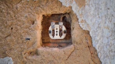 Eski bir kil duvarda, bir duvar prizinin çıplak iç kısımlarına yakın çekim. Parçalanmış bir elektrik prizinin seramik gövdesi. Kırsal bir evde eski elektrik kabloları. Pürüzsüz kamera hareketi. Yüksek kalite FullHD