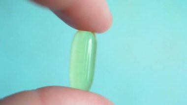 İki parmak, bulanık mavi bir arkaplanda balık yağı kapsülünü tutuyor. Omega 3 ve Omega 3 'ün kaynağı doymamış yağ asitleri. Elinde hareketli bir jelatin kabuk içinde şeffaf tablet. Yüksek