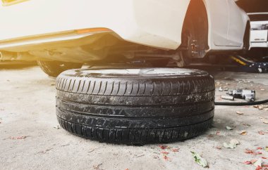 Araba tamirhanesinde arka planda tekerleği olmayan kırık bir arabayla arabanın yedek lastiği yere konmuş.