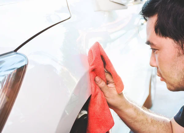 Asyalı bir adamın arabası var. Kırmızı bir bezle araba boyası silip temizliyor.