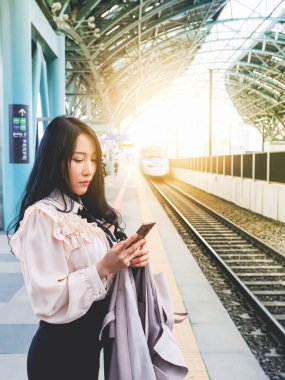 Güzel bir Asyalı kadın, tren istasyonunda trenin gelmesini beklerken akıllı telefonuyla oynuyor.