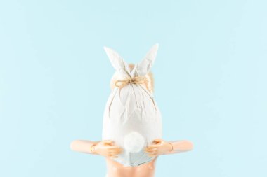 Paskalya yumurtası tutan oyuncak bebek pastel mavi arka planda tavşan şeklinde bir kağıda sarılmış. Paskalya asgari konsepti. Yaratıcı Mutlu Paskalyalar veya Bahar Düzenlemesi.