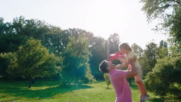 Feliz padre joven jugando con su linda hija pequeña toma a la niña en las manos y empujando hacia arriba y hacia abajo en el centro del parque junto con el resto de los miembros de la familia. Disparo en ARRI Alexa — Vídeo de stock