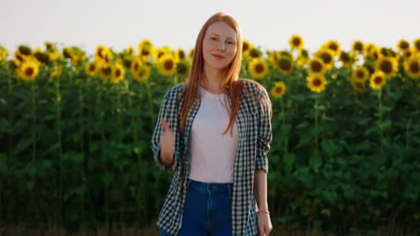 Młoda, lisiasta kobieta nosi w kratkę bluzkę i stoi przed polem słonecznikowym, pokazując kciuki do góry, uśmiechając się szeroko do kamery. Strzał na ARRI Alexa Mini. — Wideo stockowe
